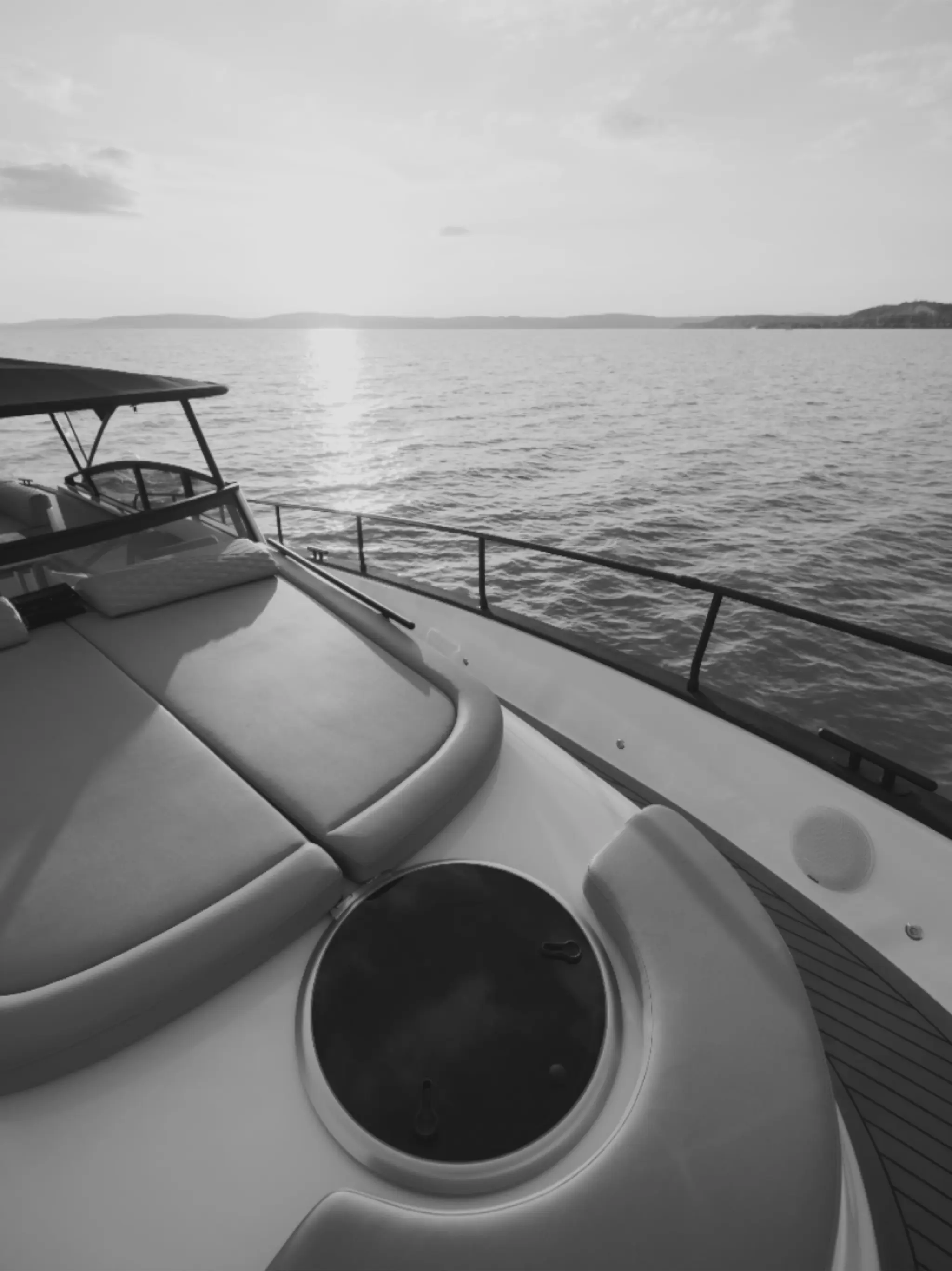 Black-and-white view across the bow sunpad of a Wia yacht towards the low sun on the water.