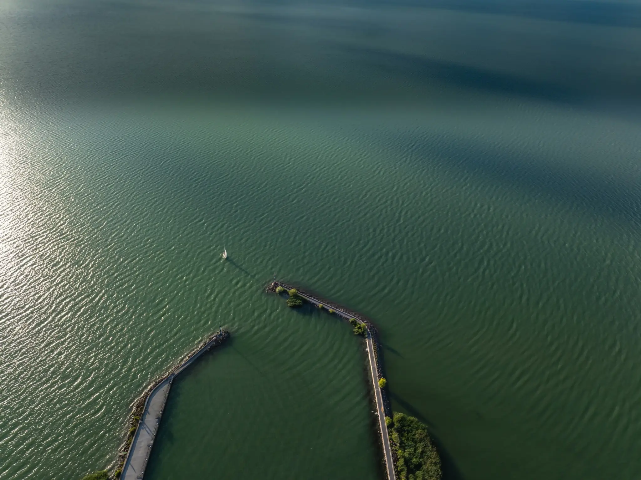 Aerial view of two stone breakwaters reaching into Lake Balaton, a lone sailboat between them.