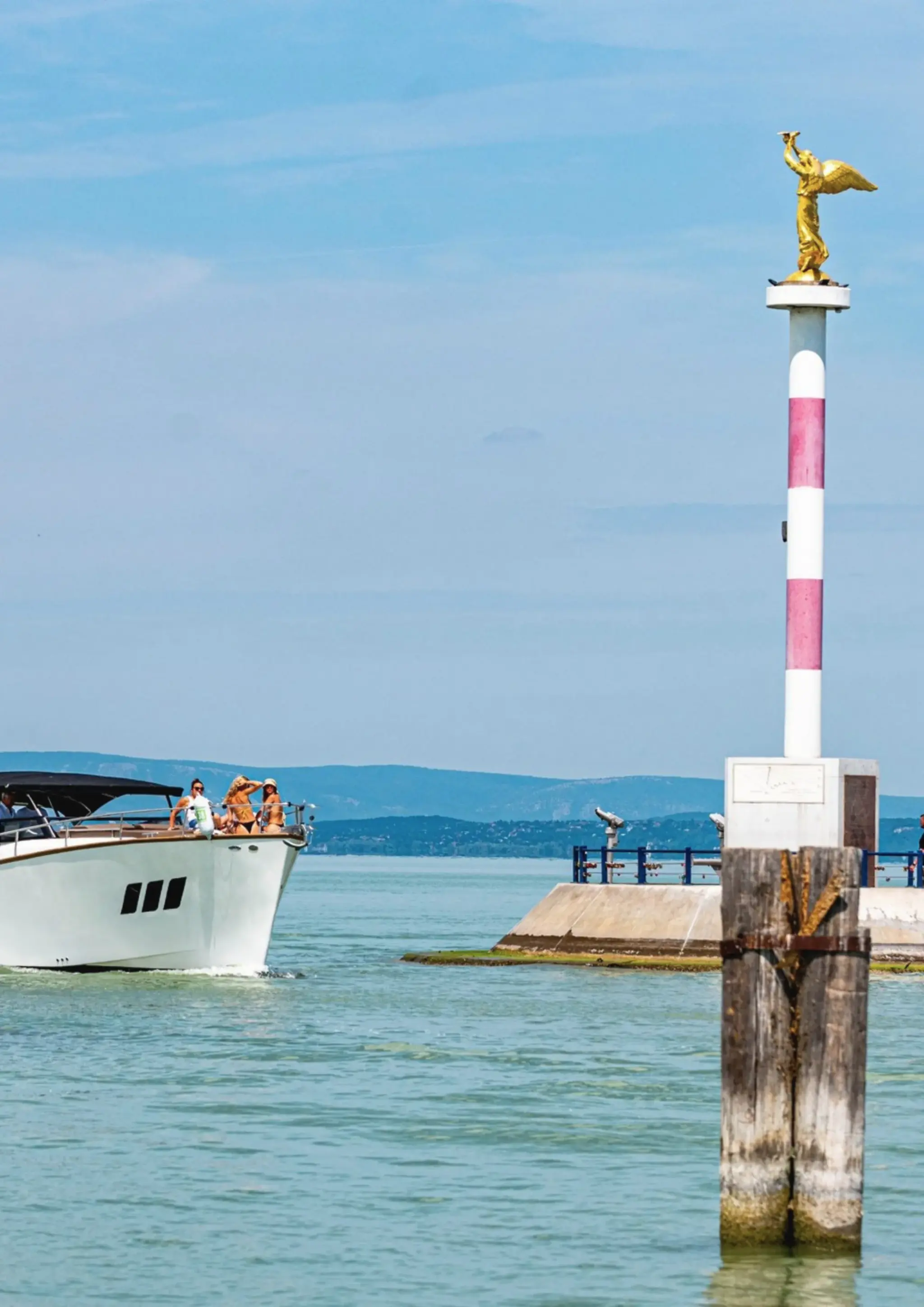 A white Wia with guests aboard cruising past a harbour entrance marked by a golden statue on a column.