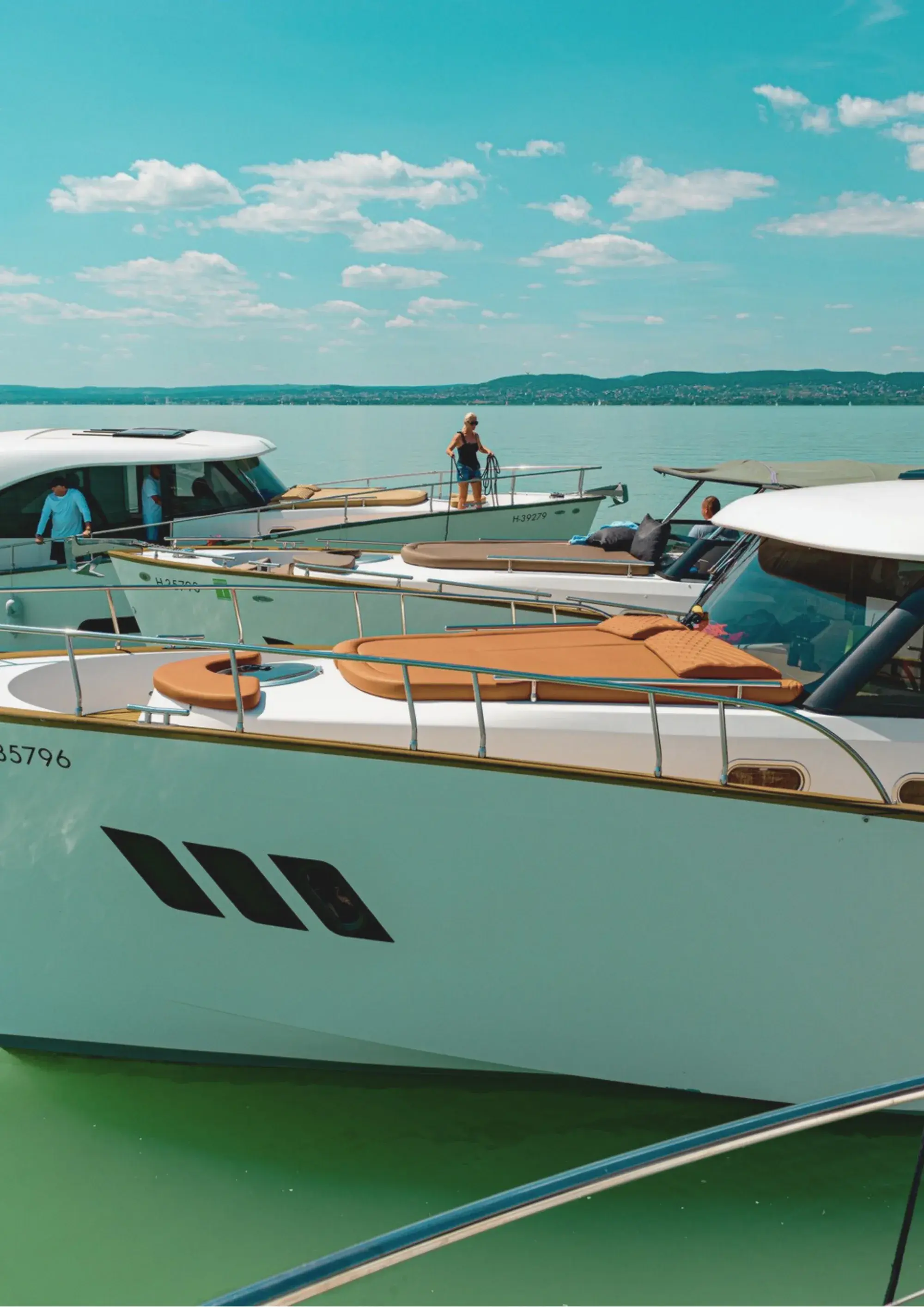 Plusieurs yachts Wia blancs aux bains de soleil beiges amarrés côte à côte dans une marina, une femme manœuvrant une amarre à la proue, les collines de l'autre rive en arrière-plan.