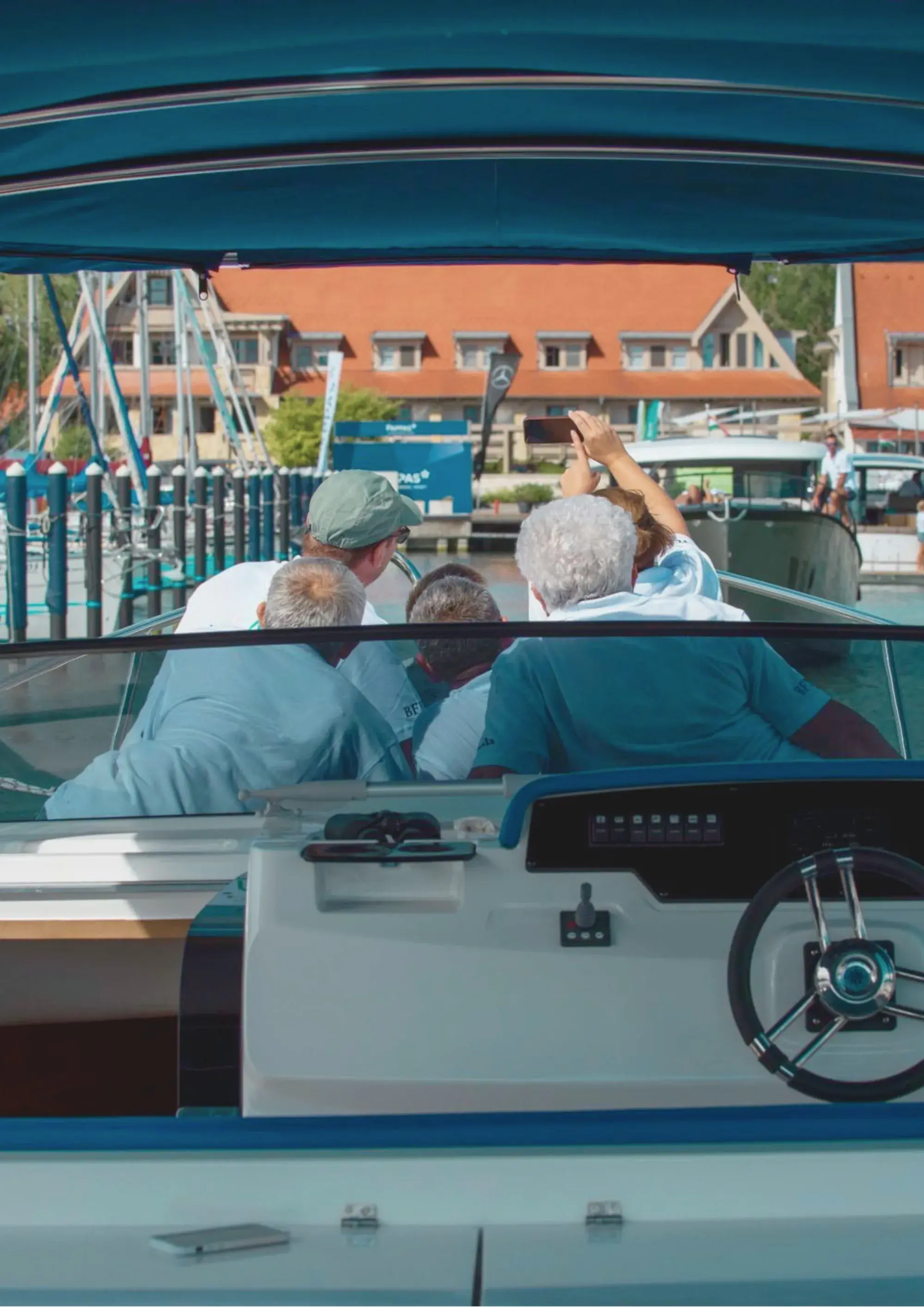 Vue vers l'avant depuis le cockpit d'un Wia sous un bimini bleu, plusieurs invités à bord dont l'un prend une photo, bâtiments de la marina en arrière-plan.