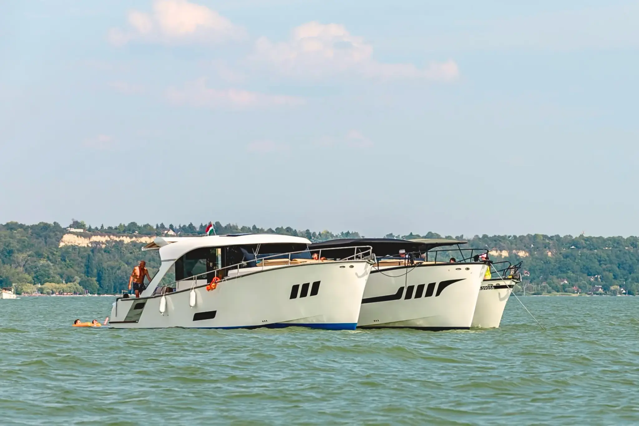 Deux yachts Wia blancs côte à côte sur le lac Balaton, un homme sur le pont latéral et deux personnes sur un matelas orange, drapeau hongrois au vent.