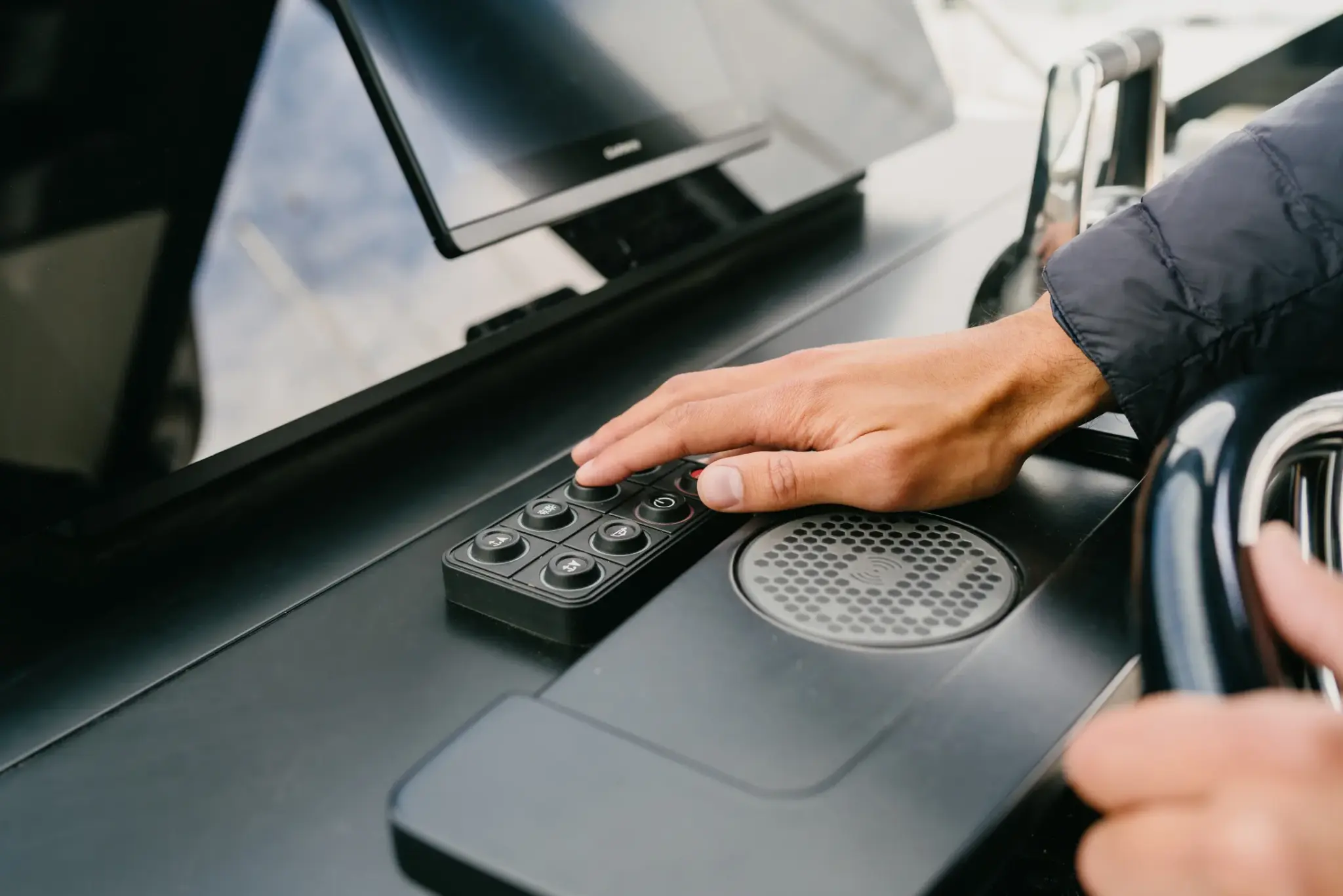 Kéz egy fekete kormányállás-kezelőpanelen, mellette Garmin kijelző és vezeték nélküli töltő.
