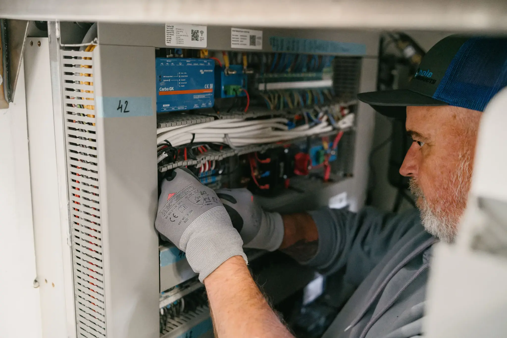 Technikus dolgozik a hajó elektromos rendszerén, kesztyűs kéz a Cerbo GX vezérlő mögötti kábelezésnél.